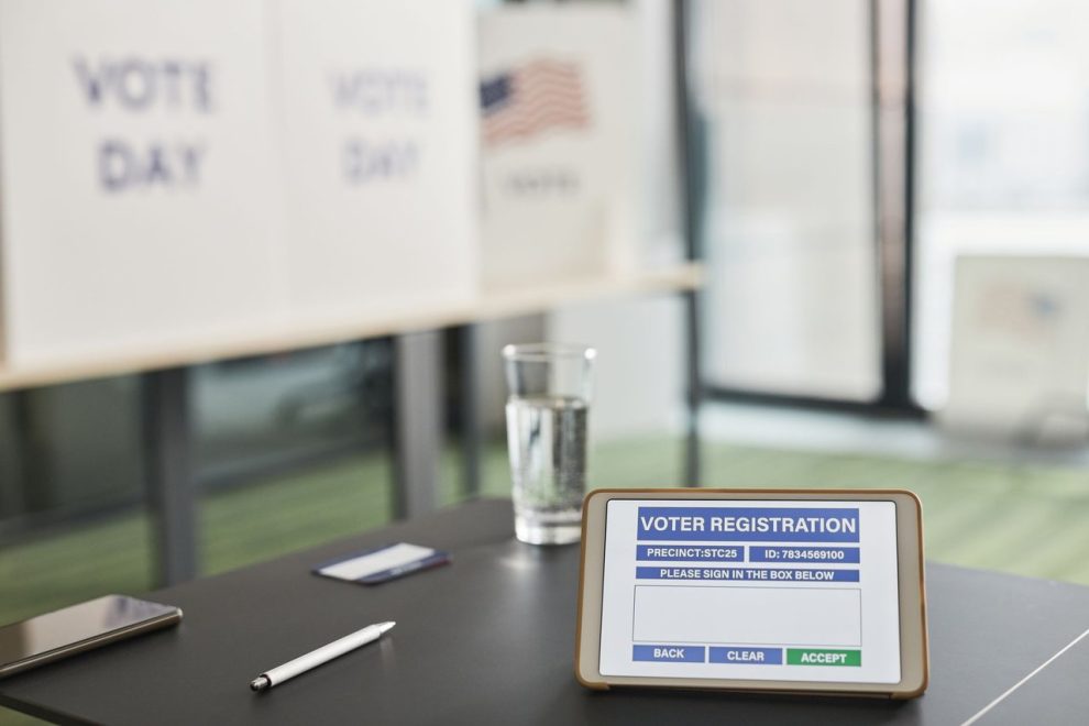 Tablet exibindo um cadastro eleitoral em uma estação de votação interna, com sinalização sobre o dia da votação.